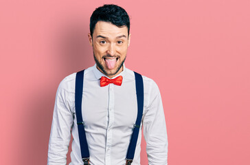 Hispanic man with beard wearing hipster look with bow tie and suspenders sticking tongue out happy...