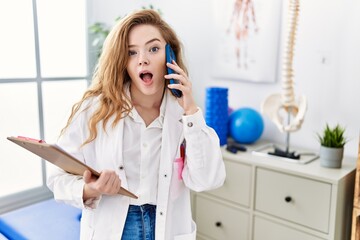Young caucasian woman working at rehabilitation clinic speaking on the phone afraid and shocked with surprise and amazed expression, fear and excited face.
