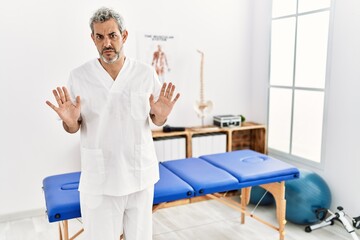 Middle age hispanic therapist man working at pain recovery clinic moving away hands palms showing refusal and denial with afraid and disgusting expression. stop and forbidden.