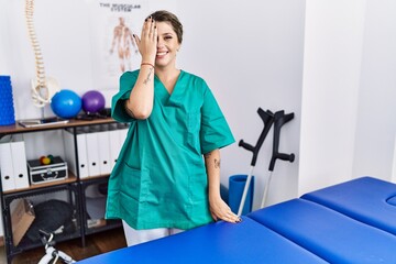 Young hispanic woman wearing physiotherapist uniform standing at clinic covering one eye with hand, confident smile on face and surprise emotion.