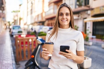 Young blonde woman using smartphone and drinking coffee at street