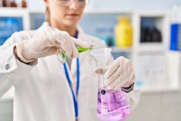 Young blonde woman wearing scientist uniform pouring liquid on test tube at laboratory