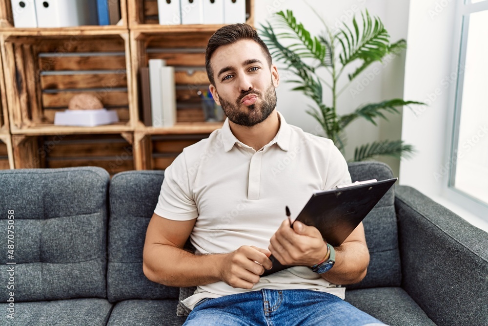 Poster handsome hispanic man holding clipboard working at psychology clinic looking at the camera blowing a