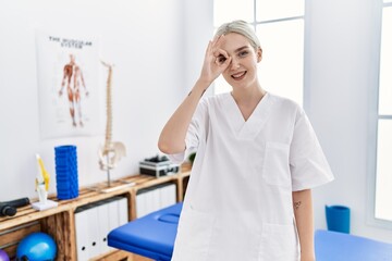 Young caucasian woman working at pain recovery clinic doing ok gesture with hand smiling, eye looking through fingers with happy face.