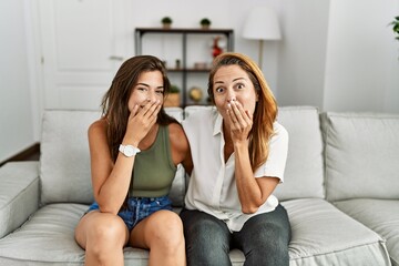 Mother and daughter together sitting on the sofa at home laughing and embarrassed giggle covering...
