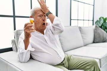 Senior man holding pills surprised with hand on head for mistake, remember error. forgot, bad memory concept.