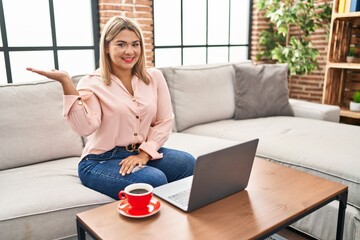 Young hispanic woman using laptop sitting on the sofa at home smiling cheerful presenting and pointing with palm of hand looking at the camera.