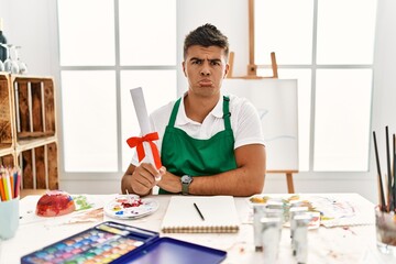 Young hispanic man at art studio holding degree depressed and worry for distress, crying angry and afraid. sad expression.