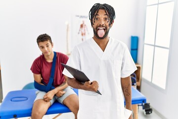 Young hispanic man working at pain recovery clinic with a man with broken arm sticking tongue out happy with funny expression. emotion concept.