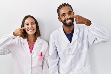 Young hispanic doctors standing over white background pointing with hand finger to face and nose, smiling cheerful. beauty concept