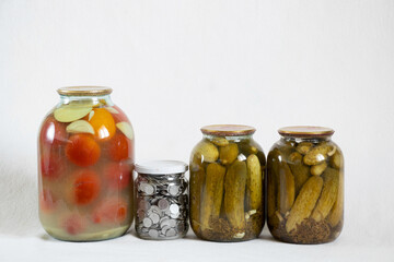 canned tomatoes and cucumbers in jars and next to a bank with Ukrainian kopecks on a white background, save money in a bank, finance