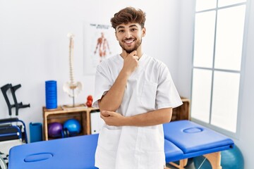 Young arab man working at pain recovery clinic looking confident at the camera smiling with crossed...