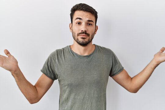 Young hispanic man with beard wearing casual t shirt over white background clueless and confused expression with arms and hands raised. doubt concept.