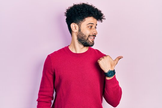 Young arab man with beard wearing casual pink sweater smiling with happy face looking and pointing to the side with thumb up.