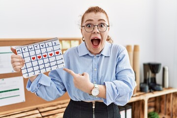 Young redhead woman holding heart calendar at the office smiling happy pointing with hand and finger