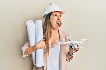 Young blonde woman wearing safety helmet holding blueprints and drone angry and mad screaming...