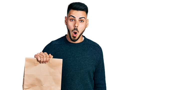 Young hispanic man with beard holding take away paper bag scared and amazed with open mouth for surprise, disbelief face