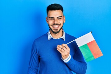 Young hispanic man with beard holding madagascar flag looking positive and happy standing and...