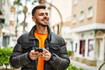 Handsome hispanic man with beard smiling happy and confident at the city wearing winter coat using smartphone