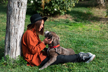 Naklejka na ściany i meble Una linda y alegre mujer caucásica con un sombrero está sentada en el parque, junto a su perro de la raza American Stafford Terrier