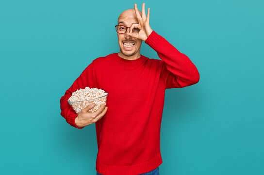 Bald man with beard eating popcorn smiling happy doing ok sign with hand on eye looking through fingers