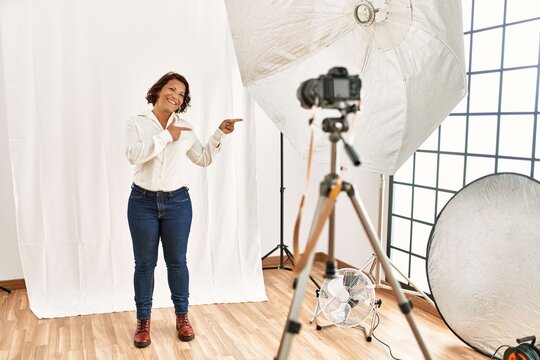 Middle age hispanic woman posing as model at photography studio smiling and looking at the camera pointing with two hands and fingers to the side.