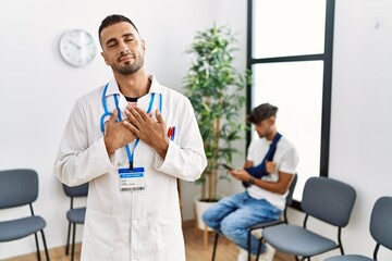 Naklejka na ściany i meble Hispanic doctor man at waiting room with pacient with arm injury smiling with hands on chest with closed eyes and grateful gesture on face. health concept.