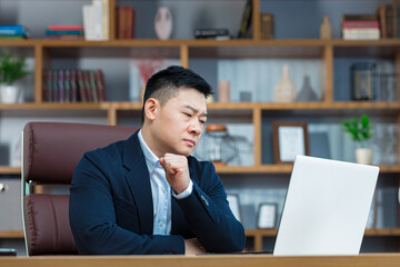 Pensive and anxious businessman, in a classic office, works at a desk with a laptop, an Asian makes...