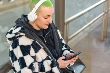 young lesbian woman waiting at the bus stop, choosing and listening music with modern devices,...
