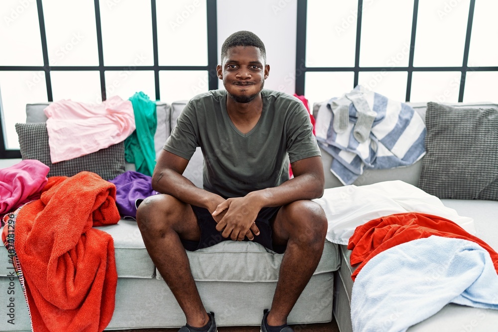 Sticker Young african american man sitting on the sofa with dirty laundry clothes puffing cheeks with funny face. mouth inflated with air, crazy expression.