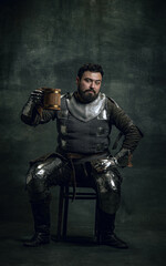 Fototapeta na wymiar Happy medieval warrior or knight with dirty wounded face holding big mug of beer isolated over dark vintage background. Comparison of eras, history