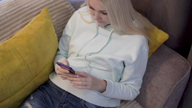 Young woman using smart phone surfing social media, checking news, doing online shooping or texting messages sitting on sofa. Spending time at home with cell gadget technology. Top-down view.