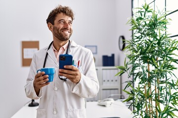 Young hispanic man wearing doctor uniform using smartphone and drinking coffee at clinic