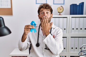 Young hispanic doctor man holding blue ribbon covering mouth with hand, shocked and afraid for mistake. surprised expression
