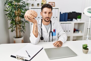 Young doctor holding brain at medical clinic thinking attitude and sober expression looking self confident