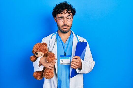 Young hispanic man wearing doctor uniform holding teddy bear and clipboard skeptic and nervous, frowning upset because of problem. negative person.