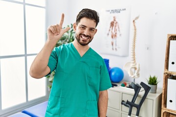 Young man with beard working at pain recovery clinic showing and pointing up with fingers number two while smiling confident and happy.