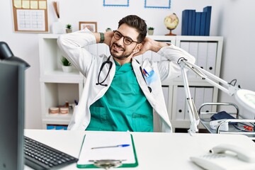 Young man with beard wearing doctor uniform and stethoscope at the clinic relaxing and stretching, arms and hands behind head and neck smiling happy