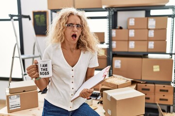 Middle age blonde woman working at small business drinking coffee cup in shock face, looking...