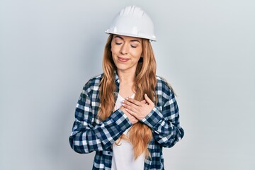 Young caucasian woman wearing architect hardhat smiling with hands on chest with closed eyes and grateful gesture on face. health concept.