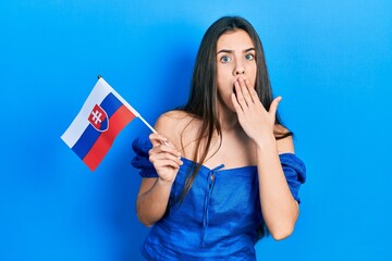 Young brunette teenager holding slovakia flag covering mouth with hand, shocked and afraid for...