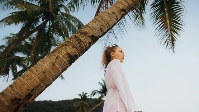 Happy woman in a white shirt on beach, near stormy sea, tilted palm tree. Attractive girl with wet curly hair in sunglasses. Female tourist walks has fun rest. Camera follow, spin rotate around model