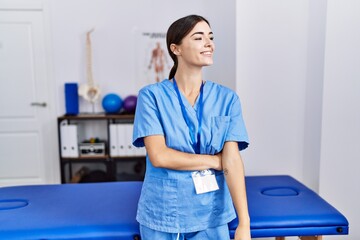 Young hispanic woman wearing physiotherapist uniform standing at clinic looking away to side with...