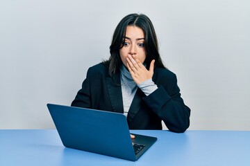 Beautiful hispanic woman working at the office with laptop covering mouth with hand, shocked and...