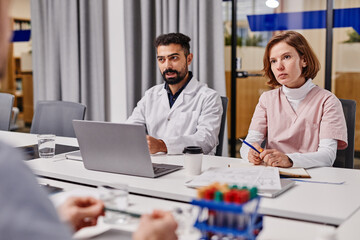 Young serious assistant in uniform and confident doctor in lab coat listening to colleagues during medical discussion at concilium