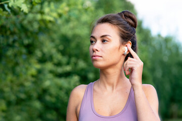 Close-up portrait of a beautiful young, pensive woman talking through bluetooth headphones while walking in the park. Serious girl, listens to music in bluetooth wireless headphones, in summer.