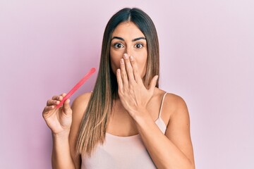 Young hispanic woman using file nail covering mouth with hand, shocked and afraid for mistake....