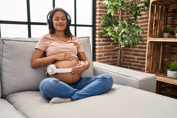 Young latin woman pregnant listening to music put headphones on belly at home - Powered by Adobe