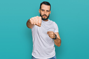 Young man with beard wearing casual white t shirt punching fist to fight, aggressive and angry...