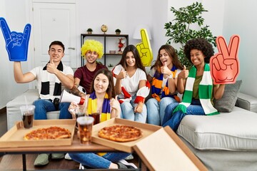 Group of young people wearing team scarf cheering football game smiling happy and positive, thumb up doing excellent and approval sign